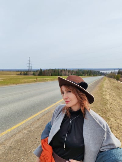 A woman is standing on the side of a road in Нечкино, Russia. She is wearing a brown hat, a gray coat, and a black shirt with a long silver necklace. She is looking to the left of the frame. Behind her is a road with a yellow line. Beyond the road, there is a field and a forest. In the distance, there is a power line tower. The sky is overcast.