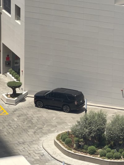 The image shows a black SUV parked on a paved area next to a modern building. The building has a light gray, textured facade with horizontal lines. There are several rectangular windows visible on the building. To the left of the SUV, there is a triangular planter with a topiaried tree and a row of small, manicured bushes. The ground is paved with light gray rectangular stones laid in a patterned fashion. In the foreground, a curved garden bed contains olive trees and various smaller shrubs. The scene appears to be an outdoor parking or entrance area of a commercial or residential building in Lusail, Qatar, bathed in daylight. There are no people visible in the image.