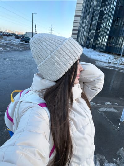 The image presents a close-up, side-profile view of a female individual in an outdoor urban environment identified as Novosaratovka, Russia. The individual, positioned off-center to the right, wears a cream-colored ribbed knit beanie and a matching cream-colored puffy winter jacket. Long, dark brown hair cascades from under the beanie and over her right shoulder. A backpack with visible yellow and magenta straps is worn over the jacket. Her face is shown in profile with a slight upward gaze and a subtle smile.
The background features a contemporary residential area under a clear blue sky. To the left, a snow-dusted parking lot contains several parked vehicles, including dark sedans and a white utility van. Utility poles and power lines are visible in the distant background. To the right and behind the individual, a multi-story modern residential building with dark, reflective window panels and a light-toned facade stands prominently. The ground in the foreground and mid-ground consists of dark asphalt partially covered by scattered patches of snow and ice, with a noticeable puddle of water on the left side. A black street lamp post is situated in the mid-ground on the left. The scene depicts a clear, cold daylight setting, indicative of winter or early spring conditions.