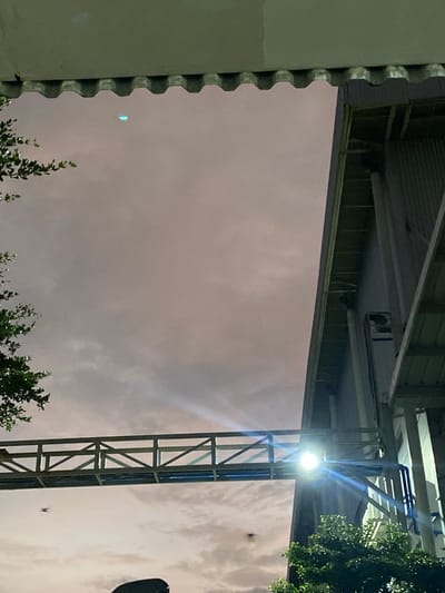 An outdoor, low-angle, wide shot captures a walkway bridge extending from a factory structure against a cloudy, twilight sky. The walkway is illuminated by a bright light fixture positioned on its side. To the left of the bridge, lush green foliage drapes downwards. The factory building, with its corrugated metal siding, dominates the right side of the frame. A small, blue, disc-shaped object is visible in the upper portion of the sky. Another dark, indistinct object appears to be in flight near the bottom left of the bridge. The overall scene is set in the city of Gambut Barat, Indonesia.
