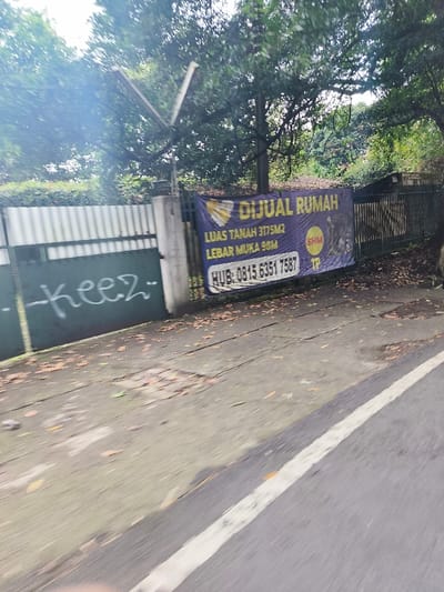 The image shows a street in Lembang, Indonesia (latitude: -6.84, longitude: 107.598). A sign on the side of the road reads “DIJUAL RUMAH” which translates to “House for Sale.” The sign is purple and yellow with a heart-shaped logo. The sign includes information about the property’s size. There is a black metal fence, graffiti on the side of the fence, and a sidewalk with leaves on it. The street is paved with asphalt and has a white line down the center. There is a black light pole visible in the image. The road is narrow and appears to be a residential street. There are trees on the side of the road, but no people are visible.