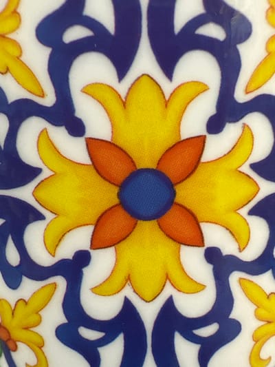 The image is a close-up of a decorative tile with a floral pattern. The pattern features a central blue circle surrounded by four orange petal-like shapes, which are in turn surrounded by four yellow petal-like shapes. These elements are outlined in dark blue and set against a white background. The surrounding area of the tile shows abstract blue swirls and smaller yellow floral elements, also outlined in dark blue. The texture of the image suggests it is printed on fabric or a textured ceramic. There is no discernible location in Coquitlam, Canada, as the image is a detail of a decorative object and does not depict any specific environment or landmarks.