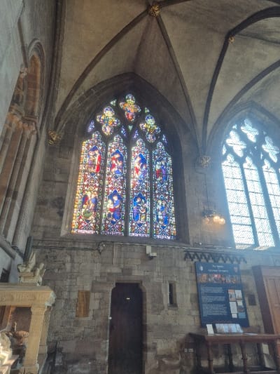 The image presents an interior view of Hereford Cathedral in Hereford, United Kingdom, characterized by its Gothic architectural style. A prominent, large arched stained-glass window, composed of multiple panels depicting religious figures and narratives in red, blue, green, and yellow tones, occupies the upper-center, illuminated by external light. To its right, a second large, arched window appears to contain plain, leaded glass, brightly overexposed by sunlight. The ceiling above consists of light grey, ribbed stone vaulting with gilded bosses at the intersections of the ribs. The lower and mid-level walls are constructed from light brown, rough-hewn stone blocks. Centrally beneath the stained-glass window is a dark wooden, arched doorway. To the left of this doorway, two small, rectangular plaques are affixed to the stone wall. To the right, a large blue informational display board titled "The Mappa Mundi & Chained Library" is mounted, beneath which rests a plain wooden table. A multi-bulb light fixture is visible on the wall near the plain window. On the far left, a carved stone monument or architectural structure with a column or pilaster is partially visible. No individuals are present in the scene.