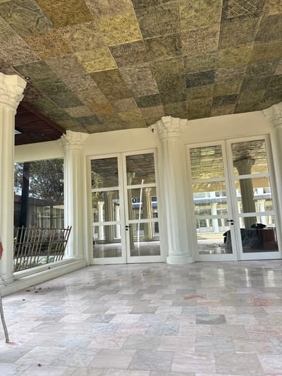 The image depicts an outdoor patio or covered walkway with a marble tile floor and a decorative metal ceiling. Large white pillars with ornate capitals support the structure. To the left, a metal fence is visible, with foliage and trees seen through a large glass window. In the center and right of the image, there are several glass doors leading into an interior space. Reflections in the glass doors show more pillars and architectural elements, suggesting a larger building or complex. The ceiling is composed of square tiles with intricate embossed patterns, varying in color from golden yellow to dark green and brown, giving it a somewhat aged appearance. The overall setting appears to be a well-maintained, possibly historical or luxurious, outdoor architectural space. The specific location of Tampaksiring, Indonesia, is not directly evident from the visual information in the image itself.