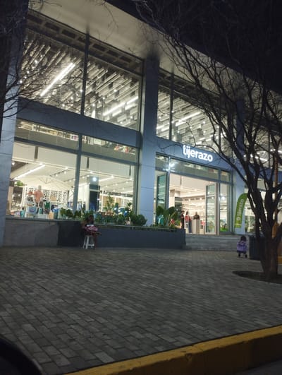 The image depicts the exterior of a "tijerazo" store at night. The building has a modern design with large glass windows showcasing the brightly lit interior. Inside, mannequins and store displays are visible. Two people are seated outside. One person sits on a small stool near the store entrance. Another person in purple clothing is seated in the distance. A tree with bare branches is visible on the right side of the frame. The ground is paved with interlocking stones, and a yellow curb is at the bottom of the frame. Based on the "tijerazo" signage, the location is likely in Valencia, Venezuela, where this retail chain operates.