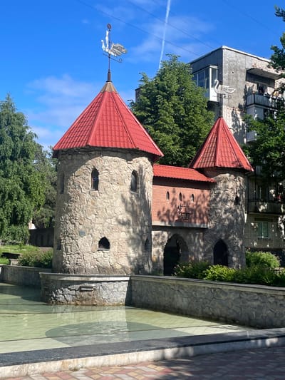 This is a vertical, eye-level, medium shot of a whimsical castle-like structure in Sosnoviy Bor, Russia. The structure features two conical red-tiled roofs and walls made of rough-hewn stone, with several small, dark arched windows. A weather vane shaped like a figure on a horse is mounted on the tip of the left turret, facing left. The castle appears to be part of a park or public space, as indicated by the adjacent paved walkway with a checkerboard pattern of pink and beige tiles, and a shallow, green-tiled pool or fountain in the foreground.

Behind the castle, an older, utilitarian apartment building with a gray facade and balconies is visible, partially obscured by lush green trees. The sky is a clear, bright blue with a few wispy white clouds and a faint contrail from an airplane. The lighting suggests it is daytime, likely midday or early afternoon, given the bright sunlight and distinct shadows.

There is no visible text in the image. The scene evokes a sense of playfulness and perhaps a touch of fairy tale fantasy, contrasting with the more mundane backdrop of the apartment building. The overall impression is a charming and unusual architectural feature set within an urban environment.