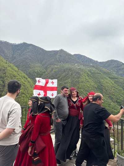 The image shows a group of people posing for a photo with the Georgian flag. The location is identifiable as the city of Kveda Chkhutuneti, Georgia, based on the mountainous backdrop. The people are wearing traditional Georgian clothing. Some are looking at the camera and smiling, while one person is taking a selfie. The scene is set on an elevated platform with a railing, overlooking the forested landscape. The weather appears to be overcast.