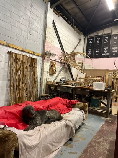 An unconventional space exhibits a mix of art and utility. Banners with the text "THEY'RE CALLING EARTH", "THEN CAME THE PLAGUE", "BUSINESS AS USUAL IS UNACCEPTABLE" are visible. A workshop area with tools and materials dominates, featuring a wooden structure, possibly an artistic installation. A dog lies still atop a red sheet. The architectural style is industrial, with exposed beams and a high ceiling. Bamboo and other crafted pieces are arranged in the space, blending the artistic with the practical.