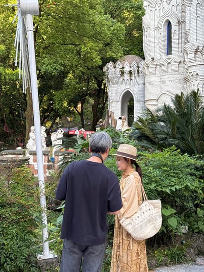 A daytime shot captures a scene near the "Fantasy Castle" in Baqiao, China. Two people are the main subjects. A woman in a light-colored dress and straw hat stands beside a man wearing a dark shirt and gray jeans. They appear to be looking at something in the distance. A white, fanciful building resembling a castle or cathedral is visible. Green trees and other greenery are prevalent throughout the scene. A white lighting structure is partially visible.