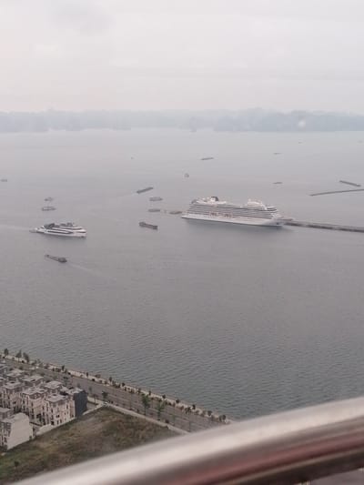 The image depicts a seascape in Hong Gai Ward, Vietnam. Several ships are visible on the water, including two large cruise ships, one docked along a pier. Smaller boats and barges are scattered across the water. A row of buildings lines the shore in the foreground, with a road and palm trees separating them from the water. The sky is overcast, creating a hazy atmosphere.