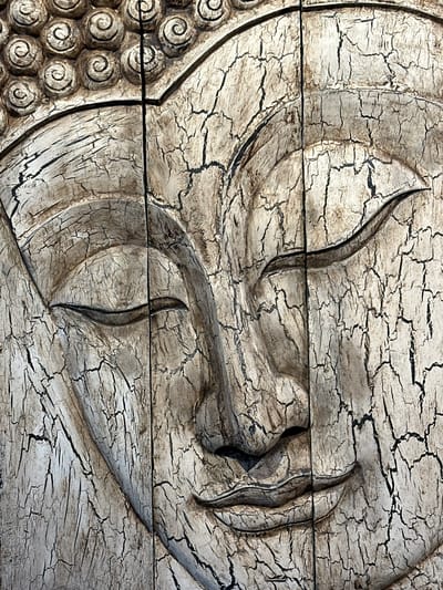 The image is a close-up, textured photograph of a carved wooden relief of a Buddha's face. The wood is heavily cracked, with a light-colored finish that reveals darker cracks and crevices, creating a distressed aesthetic. The carving depicts closed eyes, a serene expression, and the distinct coiffure of curls on the Buddha's head, which is partially visible at the top. Vertical seams in the wood are visible, dividing the face.

This image is not located in Coquitlam, Canada. It is a general depiction of a Buddha carving, not a specific landmark or scene within that city.