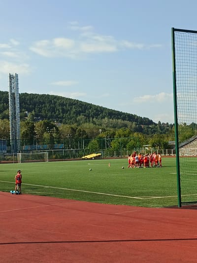 The image depicts a wide shot of a green, artificial turf soccer field in Zlatoust, Russia, under a bright, slightly cloudy sky. In the foreground, a red running track curves into view. On the soccer field, a young person in a red shirt and shorts stands on a blue scooter, facing away from the camera. Further into the field, a group of about fifteen children, mostly in red and white soccer uniforms, are gathered in a huddle. A soccer ball is visible on the grass near the huddle, as well as an orange cone. In the background, a tall cell tower or transmission tower stands to the left, and beyond that, a densely forested hill with scattered houses. To the right of the frame, a green soccer net is partially visible. The overall impression is of a sports complex in an outdoor, natural setting.