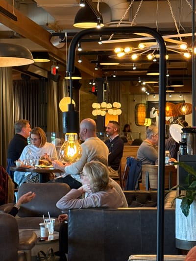 The image depicts the interior of a restaurant in Oak Brook, United States of America, focusing on multiple patrons seated at tables. Several people are seated at the tables, some are engaged in conversations, while others appear to be looking at their meals or drinks. A person is dressed in a black shirt. There is elaborate lighting present, including pendant lights with visible bulbs. The overall atmosphere suggests a lively and social dining experience. The restaurant's interior shows a mix of design elements, including wooden structures, dark furniture, and various decorative light fixtures.