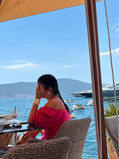 A medium shot captures a woman from the shoulders up, sitting at a table outdoors with a view of the sea in Donja Lastva, Montenegro. She is wearing a bright pink, off-the-shoulder top and has her hair in two braids. She is looking to the right, her right hand covering her mouth, and she is wearing a large gold cuff bracelet on her left wrist and a gold earring on her right ear. She is seated in a wicker chair at a dark stone table. On the table are a glass with a dark beverage and a straw, a plate, and a small lighter. The background features a calm blue sea, a distant green mountainous coastline with buildings, and several yachts anchored in the water, along with smaller boats. The scene is framed by a tan awning overhead and wooden support beams with rope details on the right. The sky is clear blue with scattered white clouds.
