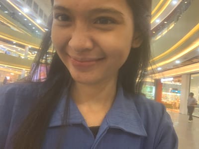 The image is a selfie taken by a person, likely female, inside a large, multi-story shopping mall in Samarinda, Indonesia. The person's face is prominent in the foreground, captured from a slightly low angle, looking towards the camera with a faint smile. She has long, dark hair framing her face and is wearing a blue top, under which a black lace bra or garment is visible.

The background displays the interior architecture of the mall, featuring multiple curvilinear levels or walkways illuminated by bright, warm yellow strip lighting and individual spotlights. The design appears modern and expansive. On the left side of the background, an inverted reflection of another person, wearing a light-colored top and dark trousers, is visible in what appears to be a glass panel or window. The lighting within the mall is entirely artificial, providing no clues about the time of day. There is no visible text in the image.