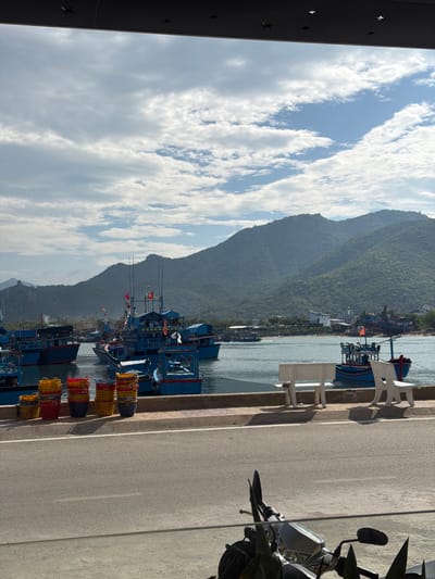 A scenic coastal view in Ninh Chu Ward, Vietnam, captures a harbor nestled between lush mountains and a partly cloudy sky. In the foreground, a paved path or road runs along the waterfront. A dark scooter is partially visible on the far right. Several white plastic chairs are arranged beside the water, and along the path, dark buckets overflow with vibrant red and yellow woven baskets or nets, indicative of local fishing activities. A dark structure with star-shaped cutouts edges the top left.

The midground features numerous traditional fishing boats, mostly painted in shades of blue with red details, docked in the calm waters of the bay. Masts and fishing equipment are visible on the vessels, some adorned with small Vietnamese flags. Across the water, a collection of white buildings lines the distant coastline.

Dominating the background are majestic, verdant mountains, thickly covered in green vegetation, providing a dramatic natural backdrop to the coastal settlement. The sky is a mix of bright blue and scattered white clouds, suggesting clear daytime weather. The overall atmosphere is one of serene coastal industry and natural beauty. No specific human activity is depicted, but the scene implies the ebb and flow of a fishing community. No visible text is clearly legible.