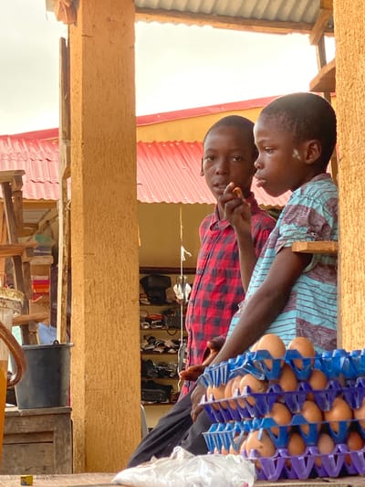 Two young Black individuals are standing adjacent to a display of eggs in Kafanchan B, Nigeria. The person in the foreground is wearing a red and black plaid shirt, while the other person is wearing a light blue and maroon striped shirt. One individual appears to be holding something in their hand. Behind them, a rack of eggs is visible. The setting seems to be an informal retail environment.