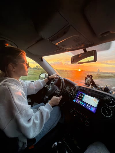 A woman drives a car during sunset. The woman is wearing glasses and a light-colored hoodie. She is looking at the road ahead. The sun is low in the sky, casting a warm glow on the landscape. The car's dashboard is visible, with a large infotainment screen displaying navigation. The interior of the car is dimly lit, with the exterior scene providing the primary light source. The location is not specified within the image or accompanying text.