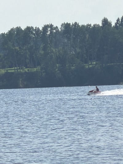 The image shows a wide body of blue water with ripples. In the middle distance, a person is riding a red and white jet ski, leaving a white wake. In the background, there is a green, tree-covered embankment. The sky is overcast and light gray. There is no identifiable landmarks to confirm the location in Saulkalne, Latvia.