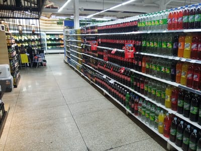 The image shows the interior of a supermarket aisle in El Valle del Espíritu Santo, Venezuela, stocked with a wide variety of bottled beverages. The shelving units are filled with numerous bottles of soda, water, and other drinks, predominantly Coca-Cola and various flavors of carbonated beverages. The aisle is well-lit by overhead fluorescent lights, illuminating the colorful labels and reflections on the bottles. The floor is made of speckled terrazzo tile. To the left of the main beverage aisle, there are also shelves displaying fruits and vegetables in refrigerated units, along with other assorted grocery items. Some shopping carts are visible in the background. The scene depicts a typical retail environment focused on food and beverage sales.
