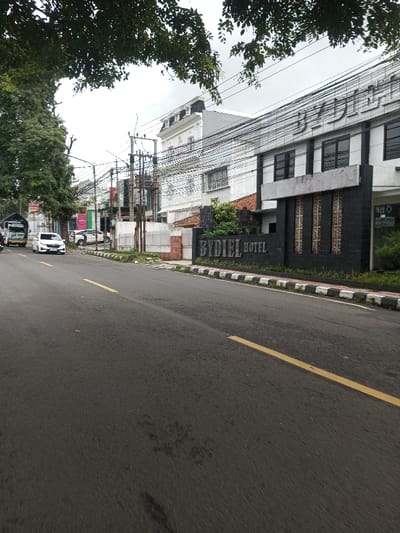 A daylight street scene is depicted in Kodarapalli, India, under an overcast sky. The view is down an asphalt road featuring two yellow dashed lines. To the right, a curb with alternating black and white stripes borders the roadway. Beyond the curb, a black sign displaying "BYDIEL HOTEL" in light-colored capital letters is present. This sign stands before the multi-story BYDIEL HOTEL building, which has a dark lower facade and lighter upper floors with dark-framed windows. Adjacent to the hotel, another multi-story white building with a prominent mansard roof is visible. The right side of the street is characterized by numerous utility poles and a dense arrangement of overhead electrical wires. On the left side of the road, a white SUV is observed, either stationary or in motion. Further down the street on the left, a light-colored truck and additional vehicles are visible. Green trees line the left side of the road. No individuals are present in the image.