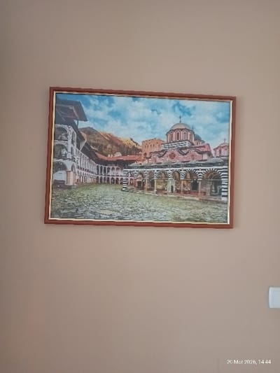The image, captured on 20 Mar 2026 at 14:44, displays a framed picture hanging on a plain, light beige wall. The scene is located in Vedrare, Bulgaria. The rectangular picture is enclosed in a brown wooden frame with a narrow golden inner trim. The artwork within the frame depicts the Rila Monastery, showcasing its central courtyard. Elements visible in the picture include a cobbled ground, multi-story monastic buildings with white facades, arched colonnades, and dark roofs. The main monastery church is prominently featured on the right side, characterized by multiple domes, including a large central one, and distinctive horizontal red and white striped banding on its exterior walls. Rolling hills or mountains are visible in the background beneath a blue sky. A portion of a white electrical fixture or switch is visible on the lower right section of the wall, outside the picture frame. There are no people, actions, or interactions depicted.