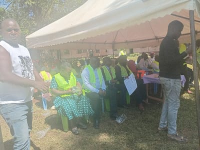 An outdoor gathering is depicted in Idakho South ward, Kenya, featuring multiple individuals under a large white canopy tent erected on a grassy area. In the foreground left, a dark-skinned male subject, wearing a white sleeveless t-shirt with a dark graphic and light blue jeans, stands facing slightly right, hands clasped. Under the tent, a seated audience occupies green and other colored plastic chairs. Many seated subjects, predominantly dark-skinned, wear bright yellow-green safety vests over various clothing. A prominent seated dark-skinned female subject, wearing a green polka-dot dress under a yellow-green vest, holds a small, clear plastic-wrapped object in her hands. Other seated male subjects in vests are visible, one wearing a light blue shirt. A white document with writing is visible near a seated individual. To the foreground right, a dark-skinned male subject, wearing a black long-sleeved shirt, light-wash jeans, and brown sandals, stands facing away from the camera, partially obscured by a tent pole. A small table draped with a light-colored tablecloth is present under the tent. The background features green foliage from trees and a light-colored building with dark windows or roof. Loose clear plastic items are scattered on the grass. The scene is brightly lit by natural daylight.