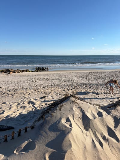 A young boy is playing on a sandy beach in Hampton Bays, New York. He is wearing blue and white patterned swim trunks and is bent over, looking at the sand. The beach is wide and sandy, with dunes in the foreground and the ocean stretching out to the horizon. The water is a deep blue, with gentle waves lapping at the shore. There are some rocks and old wooden pilings on the beach, suggesting it may be a historical site. The sky is a clear, bright blue with a few wispy clouds. The sun is casting long shadows across the sand, indicating it is late afternoon or early evening.
