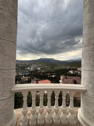 A high-angle shot captures a panoramic view of Alushta, Ukraine, under a dramatic, overcast sky. The foreground features a white balustrade and parts of two textured white columns, framing the cityscape. Buildings, varying in height and architectural style, are nestled amongst dense evergreen trees. Beyond the immediate urban sprawl, rolling hills and mountains stretch towards the horizon. The light suggests either early morning or late afternoon, with a subdued brightness breaking through the clouds. No people are visible in the image.