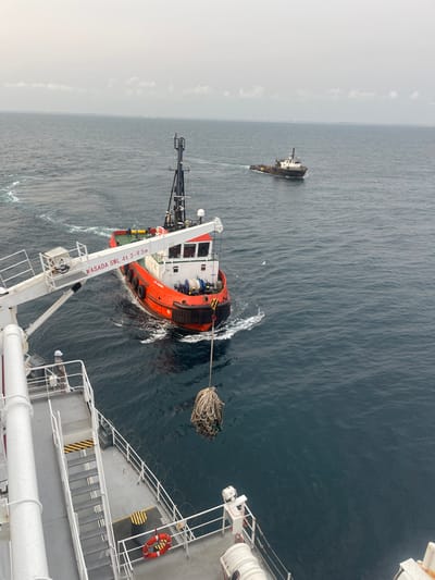 The scene depicts a maritime transfer operation occurring at sea off the coast of Orimedu, Nigeria, under an overcast sky. The image is taken from an elevated perspective on the deck of a large vessel, which features grey decking, white railings, and a prominent white crane arm extending over the water on the left. Text "NASADA SWL 4t 3-9.5m" is visible on the crane arm. From a hook at the end of this crane, a large, dark-colored bundle of tangled ropes or netting is suspended directly above the water.
On the deck of the foreground vessel, a single individual wearing a white hardhat and light-colored workwear is visible, positioned near the railings. Stairs with yellow and black hazard stripes ascend towards the upper deck on the left. In the lower right, a red and white lifebuoy is attached to the railing, which also features a line of razor wire along its top.
To the right of the foreground vessel, a red and white tugboat or workboat is positioned, creating a wake from its stern, indicating movement. This vessel has a black fendering system around its hull and a mast structure on its deck. Further in the background, a second, darker-colored vessel, also appearing to be a tug or workboat, moves away from the scene, leaving a visible wake on the dark blue-green water. The horizon is visible in the distance, meeting a hazy, light grey sky.