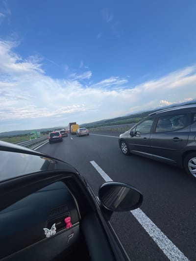 This media file depicts a traffic jam on a highway. The camera is inside a car, looking forward and slightly to the right. The scene shows multiple vehicles stopped on the road, including cars, a yellow truck, and a green truck. The sky is blue with some white clouds, suggesting it is daytime and the weather is fair. The highway has white lane markings. In the foreground, part of the interior of the car is visible, including a dashboard with a pink bow-shaped air freshener and a butterfly sticker. The surrounding landscape appears to be rural with green hills and trees. The overall impression is of a frustrating delay, common in traffic jams. The specific location is identified as Vakarel, Bulgaria. There is no visible text in the image.
