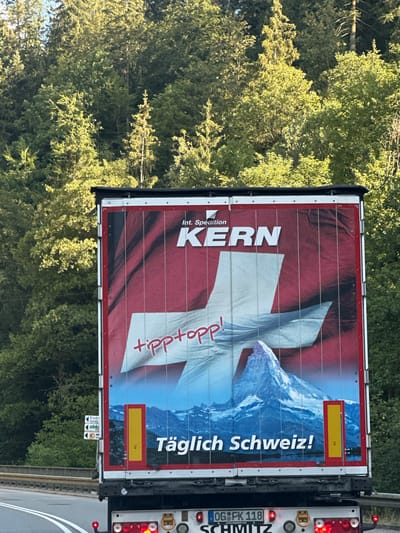 This image shows the rear of a truck trailer with a prominent advertisement. The trailer is red with white vertical stripes and features a large Swiss flag graphic. The words "Int. Spedition KERN" are printed in white at the top of the trailer, with "KERN" being significantly larger. Below this, the phrase "tipp topp!" is written in red script. In the lower portion of the trailer, a snow-capped mountain, resembling the Matterhorn, is depicted against a blue sky with a body of water and distant mountains. The German text "Täglich Schweiz!" is printed in white at the bottom. On the left side of the trailer, a small road sign with directional arrows and text like "Eisen Hans" is visible. The truck is on a road, with a dense forest of green trees forming the background. The scene appears to be in Schonach im Schwarzwald, Germany.