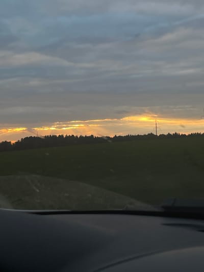 The image shows a sunset over a rural landscape. The sky is filled with dramatic clouds, with rays of sunlight breaking through in the distance. Below the sky is a dark silhouette of a forest, and in front of that, a green field. The foreground shows a dark, reflective surface, likely the dashboard of a vehicle, suggesting the photo was taken from inside a car. There are two tall, slender electrical pylons visible in the distance, one slightly to the left of center and another further to the right. The overall impression is one of natural beauty and twilight. The specific location in Povarenki, Russia is not discernible from the image itself.