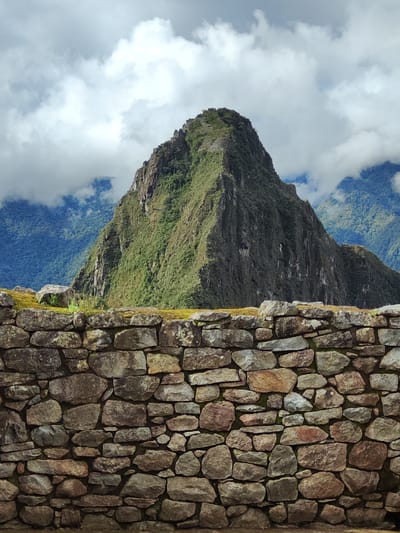 The image displays a majestic mountainous landscape, identified as Machu Picchu, Peru, dominated by a steep, verdant peak in the background. In the foreground, a meticulously crafted stone wall, typical of Inca architecture, spans the width of the frame. This wall is constructed from varied sizes of irregularly shaped, tightly fitted stones, exhibiting natural hues of grey, brown, and ochre, with patches of green moss on its upper surface.
Beyond the wall, the prominent mountain rises sharply, its slopes densely covered with lush green foliage and rugged rock formations. The cropped sections reveal what appear to be ancient terraces or small structures on the upper ridges of this peak, hinting at human modification of the natural landscape. In the far distance, additional layers of hazy, blue-green mountains recede into the backdrop.
The sky is partly cloudy, with bright white clouds interspersed with darker grey formations, suggesting a dynamic weather pattern, likely a cool or temperate climate often found in high-altitude regions. The lighting indicates it is daytime, possibly late morning or early afternoon. No human activity or discernible text is visible, emphasizing the ancient, untamed, and preserved nature of the site. The scene evokes a sense of awe for both the natural grandeur and the historical human ingenuity.