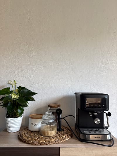 The image displays a close-up, eye-level shot of a coffee station on a wooden countertop against a textured, off-white wall. On the left, a potted white anthurium plant with dark green leaves sits in a white textured pot. To the right of the plant are two glass jars and a white sugar container. The first jar, closer to the plant, has a wooden lid and is labeled "SUGAR" in black lettering. The second jar, taller and ribbed, contains brown sugar. Next to these is a tall glass jar filled with coffee beans. A black coffee scoop rests against the coffee bean jar. To the right of the jars is a black espresso machine with a silver accent on its handle and a control panel with buttons labeled "espresso," "2x espresso," "favorite," and "steam." The machine has a chrome drip tray with the brand name "CROWN" visible. The overall scene is a neatly arranged coffee preparation area. The location is not specified in the image, and therefore cannot be determined to be Momchilovtsi, Bulgaria.