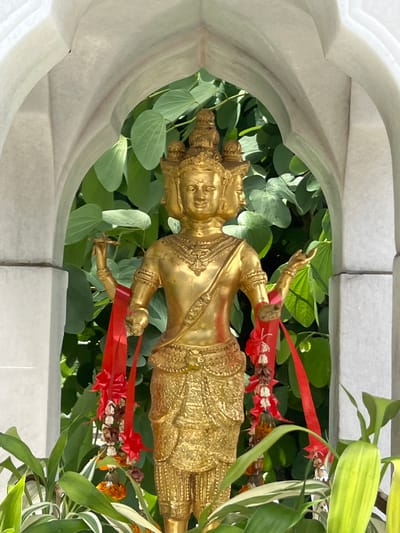 A golden statue of a multi-faced deity stands within a white arched alcove. The statue depicts a figure with four faces, each with serene expressions. The deity is adorned with intricate golden jewelry and clothing, including a crown with multiple tiers. Red ribbons and garlands with dried flowers are draped around the statue's arms and lower body. In the background, lush green foliage is visible through the arch. The statue is positioned outdoors, with green plants in the foreground. The architectural style of the alcove suggests it is part of a temple or shrine, potentially located in Pa Khlok, Thailand.