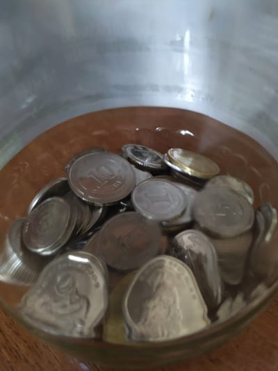The image shows a close-up, high-angle view of coins inside a glass jar, which is placed on a wooden surface. The jar is partially filled with numerous silver and gold-colored coins, many of which are stacked or overlapping. The coins vary in size and design, with some exhibiting visible markings and text.
One prominent coin in the foreground displays the number "10" within a circular design. Another coin has text that appears to be in Cyrillic script, partially visible as "ТУРКА". Other coins show partial inscriptions and national emblems. The lighting suggests an indoor setting, with the bright light reflecting off the coins and the glass jar. The overall scene conveys a sense of accumulated wealth or savings. No people are visible in the image. The location context provided is Turka, Ukraine.