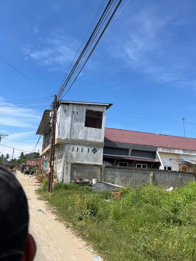 The image depicts a street scene in Dalam, Indonesia, under a clear blue sky with scattered clouds, suggesting it is daytime. The foreground is dominated by a person's head and shoulder, partially obscuring the view. A dirt road runs alongside a narrow strip of lush green grass and vegetation.

Several buildings are visible. To the left, a two-story concrete structure with a metal-barred window on the upper level stands out, with many electrical wires and a utility pole in front of it. Behind this, a larger building with a reddish-brown tiled roof and grey and white exterior is present. This building has multiple windows, some of which are dark and appear to be empty. Further down the road, more buildings can be seen, including what looks like a shop or residence with a darker facade and a small red flower bush in front. Several motorcycles are parked along the side of the road.

The overall scene appears to be a typical rural or semi-urban environment. There is no discernible activity or event taking place, other than what is implied by the presence of parked vehicles. The general atmosphere is bright and sunny, indicative of good weather. Some faint text is visible on a white sign attached to the utility pole, but it is too small and blurry to be legible.