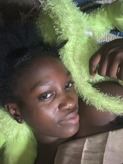 A forensic description of the scene depicts a close-up overhead view of a young individual, likely female, with dark skin and dark, braided hair, lying down. The subject's head is positioned towards the left of the frame, with her face turned slightly upwards, looking directly at the camera. Her expression is neutral. A small, dark stud earring is visible in her left earlobe.

A vibrant lime-green, highly textured, fuzzy fabric covers the right side of the subject and extends across the middle and upper right portions of the frame. A dark-skinned hand is visible on the right, resting on this green material. Portions of a blue and white patterned mat are discernible beneath the green fabric in the upper right background. A flat, brown, cardboard-like object is present in the lower right foreground. The overall lighting is subdued. This scene is observed in Agege, Nigeria.