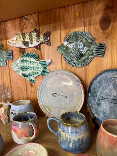 The image shows a close-up view of handcrafted ceramic items displayed on a wooden shelf. In the upper portion of the image, three ceramic fish decorations are hung on a wooden plank wall. The fish on the left is horizontally oriented with brown, green, and white stripes and a light brown belly. The middle fish is green with darker green speckles and large white eyes. The fish on the right is also green with a speckled texture and has darker green accents. Below the fish, two ceramic plates are visible. The plate on the left has a light cream-colored base with pinkish-red accents on the rim and a drawing of a fish with the inscription "Белое Море" (White Sea). The plate on the right is darker blue with a speckled pattern and a similar drawing of a fish with the same inscription. In the foreground, three ceramic mugs are visible. The mug on the left is light blue with an inscription and a drawing of birds. The middle mug has a rounded shape with a glaze that transitions from blue and grey at the top to brown and yellow at the bottom, also featuring the "Белое Море" inscription and birds. The mug on the right is a warm pinkish-orange color. The overall environment suggests a shop or display area for artisanal crafts, likely located in Solovetskiy, Russia, given the inscription on the plates and mugs.
