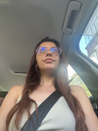 A low-angle, medium shot captures a young woman with long, dark brown hair and glasses inside a vehicle. She is wearing a light-colored, strapless top and a black seatbelt. The interior of the car, including the ceiling and a grab handle, is visible. Through the window on the right, a brick building and trees are partially visible under a bright sky, suggesting an outdoor environment. The woman is looking upwards and to the right.