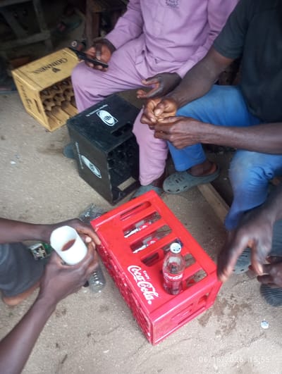 In Dokan Tofa, Nigeria, a scene unfolds outdoors amidst a sandy, gravelly ground. Four individuals are present, two of whom are in the foreground. One person is wearing a light purple traditional garment, holding a mobile phone in their right hand. Next to them, another person in a black t-shirt and blue trousers is also seated. In the foreground, a person's hands are visible holding a white cup filled with a dark liquid, and near it, a plastic bottle. A red Coca-Cola crate is prominent, containing a bottle of Coca-Cola. To the left, a light-colored wooden crate, possibly for beverages, is visible, labeled "GUINNESS." A black crate, likely for bottles, is also present, with the "NB" logo. The environment appears to be an informal setting, possibly a market or roadside stall. The timestamp "06/16/2026 15:55" is superimposed on the image.