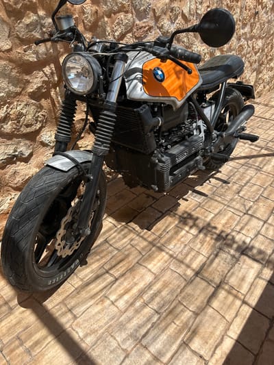 The image shows a custom BMW motorcycle parked on a stone-tiled patio in front of a textured stone wall. The motorcycle is a street scrambler style with an orange fuel tank featuring the BMW logo. It has a round headlight, exposed engine components, and knobby tires. The patio is illuminated by sunlight, casting shadows from the motorcycle and overhead foliage. The location is identified as Santa Maria del Camí, Spain. There are no people or animals visible in the image. The scene is static and appears to be taken outdoors during the day.