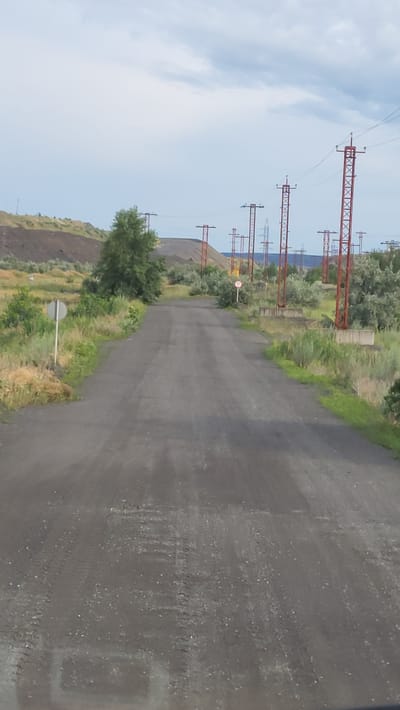 The image shows a rural road leading through a landscape with a large pile of dark material, likely industrial waste or coal, on the left. A line of red and metal electricity pylons runs along the right side of the road, extending into the distance. The road is paved and appears to be in good condition, with tire tracks visible on the surface. Lush green vegetation and scattered trees border the road and the industrial pile. The sky is overcast with some clouds visible. The scene is devoid of any people or vehicles, creating a sense of stillness and isolation. This location is identified as being in the city of Yerystivka, Ukraine.