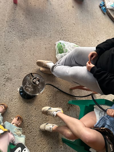 A high-angle shot shows a concrete patio area with several individuals present. In the foreground, a round, metal hookah bowl filled with charcoal sits on the ground, connected to a black hose. To the left, the feet of two individuals are visible; one person wears pink sandals with dark nail polish, and the other wears torn blue jeans. Further to the right, a person in light gray sweatpants and beige clogs is seated. Another individual, also in light gray sweatpants, is seen from the waist down, wearing a black hoodie and holding a small orange object. To the far right, a person is seated in a green chair, wearing light-colored shorts and a blue and white striped shirt, with their legs crossed. A white plastic bag rests on the ground near the seated individuals. The environment appears to be outdoors, possibly a backyard or patio. The location in the city of Пепеляева, Russia, is not evident from the visual information provided in the image.
