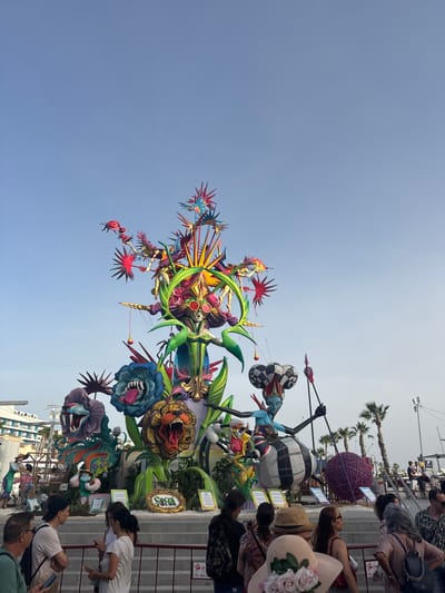 The image depicts a large, colorful monument, likely a "ninot" from the Fallas festival, in Alicante, Spain. The monument is elaborate, featuring fantastical figures, large animalistic heads with open mouths, and vibrant floral elements. A central female figure with blue skin, a jester-like hat, and elaborate clothing stands astride a large striped sphere. Smaller figures and mythical creatures are integrated into the structure. The monument is surrounded by a crowd of people, some of whom are looking at the monument, while others are engaged in conversation or looking at their phones. Palm trees and buildings are visible in the background, suggesting an outdoor urban setting. The sky is clear and blue. A low barrier with signage is in the foreground, indicating a viewing area or event space.