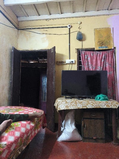 An interior room in Kochi, India, is presented with a partial view of an individual resting on a bed in the lower-left foreground. The person's bare feet and lower left leg, clad in dark trousers, are visible on a bed covered with white and red patterned bedding featuring a house-like motif. To the right, a dark flat-screen television sits on a wooden table draped with a light-colored floral tablecloth. A green object, possibly a hat or container, is on the table beside the television. Under the table, a white sack and a small, dark wooden cabinet with a single drawer are visible.
The floor is a reddish-brown. The primary wall is painted a faded light yellow, exhibiting signs of wear and discoloration, while a section to the far right is painted light purple. A dark wooden double door, partially open, reveals a darker, ambiguous space beyond. Black electrical conduit runs along the top section of the yellow wall, leading to a white switchboard with multiple outlets and switches. Above the switchboard, a white spherical device is mounted on the conduit, with a dark, teardrop-shaped object hanging below it. A yellow-framed artwork, displaying script identified as Arabic, is affixed to the wall to the right, above a window partially obscured by a red floral curtain. The ceiling is white, featuring visible beams.