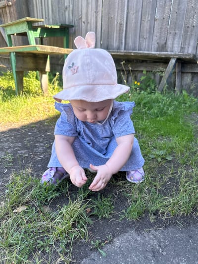 A young child wearing a blue dress and a pink sun hat with bunny ears is crouching in a grassy area. The child is looking down and appears to be playing with the grass. In the background, there is a wooden fence and some old green wooden furniture. The setting appears to be outdoors, likely in a yard. The image was taken in Novoyeniseyka, Russia.