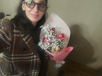 A woman wearing round glasses and a brown plaid jacket holds a bouquet of red and cream tulips interspersed with baby's breath. The flowers are wrapped in pink paper with a small tag. The background is a pale green wall.