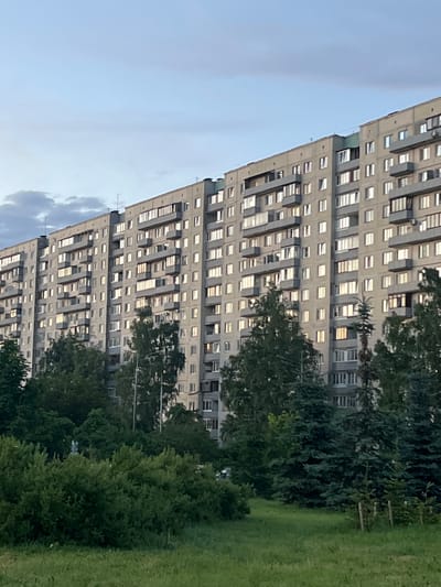 The image shows a large, multi-story apartment building in Ladozhskoe Ozero, Russia. The building is constructed of concrete panels and has a uniform design with many windows and balconies. The facade is a light grey color, and the balconies have railings. The building is surrounded by lush green trees and grass, suggesting a park-like or residential area. The sky is a pale blue with some scattered clouds. The overall impression is of a typical urban residential landscape. There are no people visible in the image.